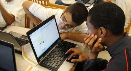 Group of diverse students and teacher smiling in classroom, wearing colorful t-shirts, sitting and standing around a green table with lockers and windows in background