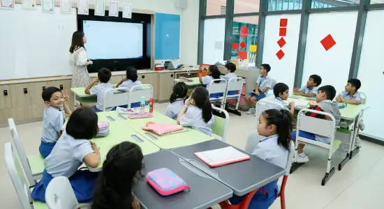 Group of diverse students and teacher smiling in classroom, wearing colorful t-shirts, sitting and standing around a green table with lockers and windows in background